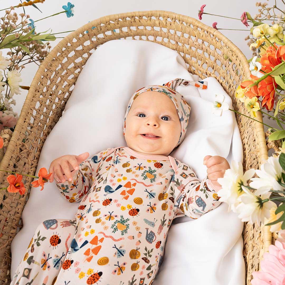 Newborn baby laying down on a cradle showcasing our convertible Garden Friends pajama. The print showcase ladybugs, butterflies, flowers, birds, snails, cactus, bees and plants. 