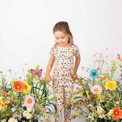 Toddler showcasing our Garden and Friends 2 Set Pajama on a white Background with flower props. The print showcase ladybugs, butterflies, flowers, birds, snails, cactus, bees and plants. 